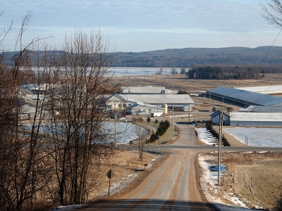 [Coming down a hill in a car the view spread out before us was of the frozen water ponds on either side of the road and multiple large buildings across the intersection with the frozen river in the background. Snow can be see only at the edges of the roads.]